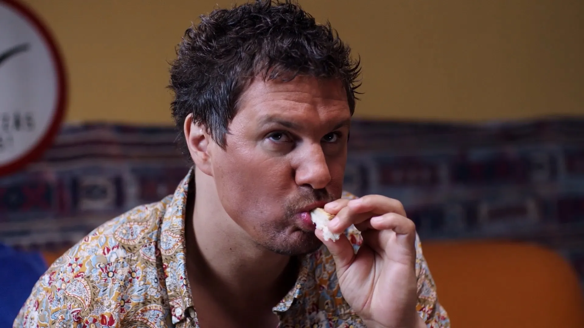 A man with tousled hair eats a piece of bread while staring upward with a slightly puzzled, absorbed expression.