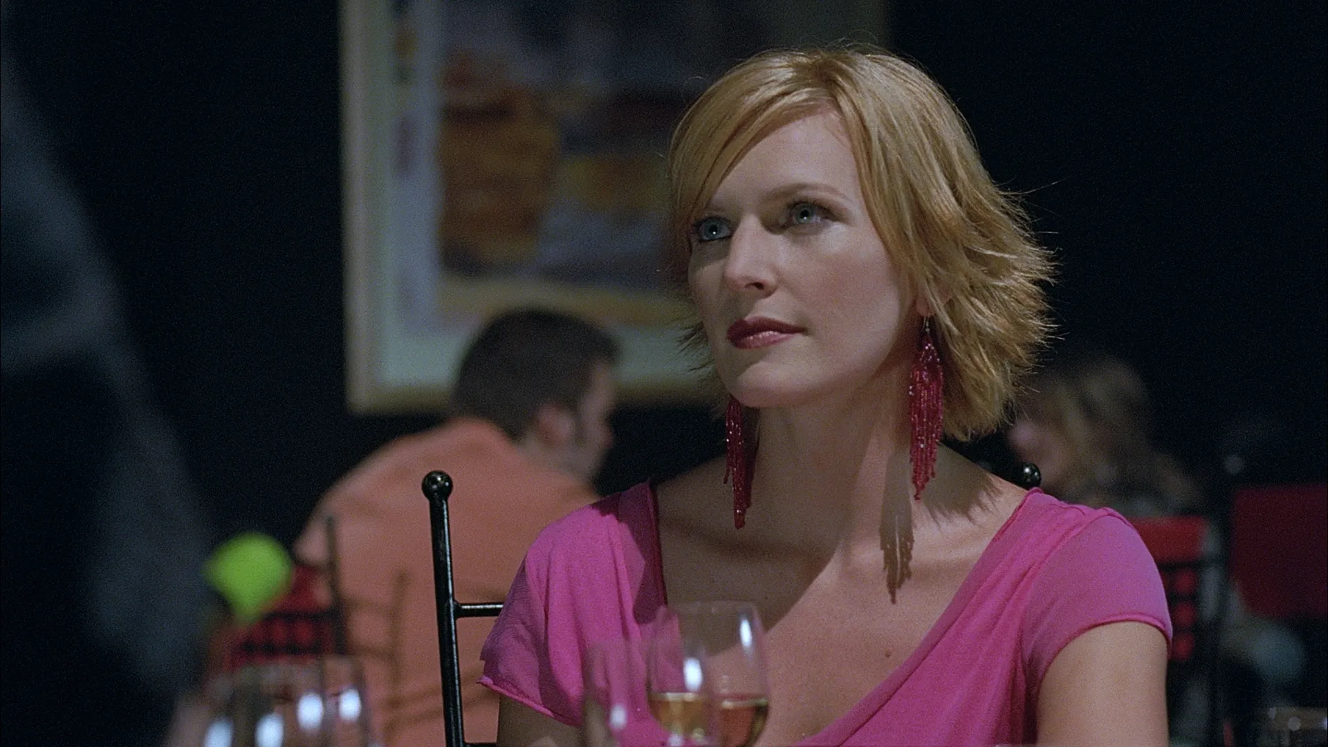 A blonde woman in a pink top and long red earrings sits at a table with a glass of white wine, her expression poised and attentive under soft, moody restaurant lighting.