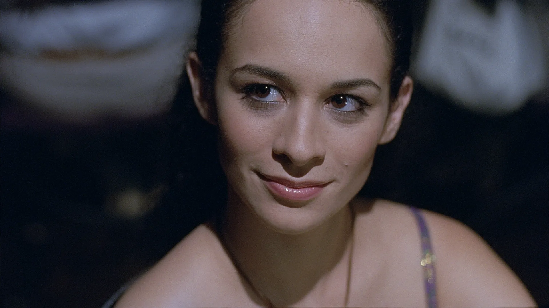 A close-up of a woman with dark hair and expressive eyes, giving a subtle, knowing smile under soft, cool-toned lighting, with the background gently blurred to keep the focus on her face.