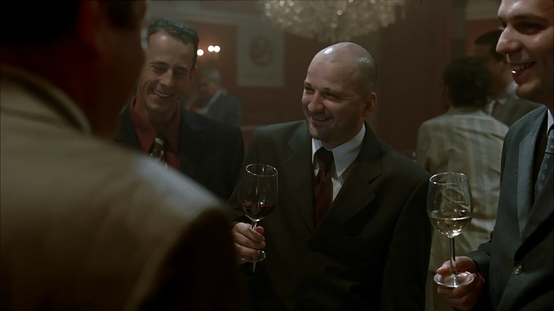 A group of well-dressed men stand together at a formal reception, smiling and holding glasses of wine as they share a lighthearted moment in the warm, softly lit room.