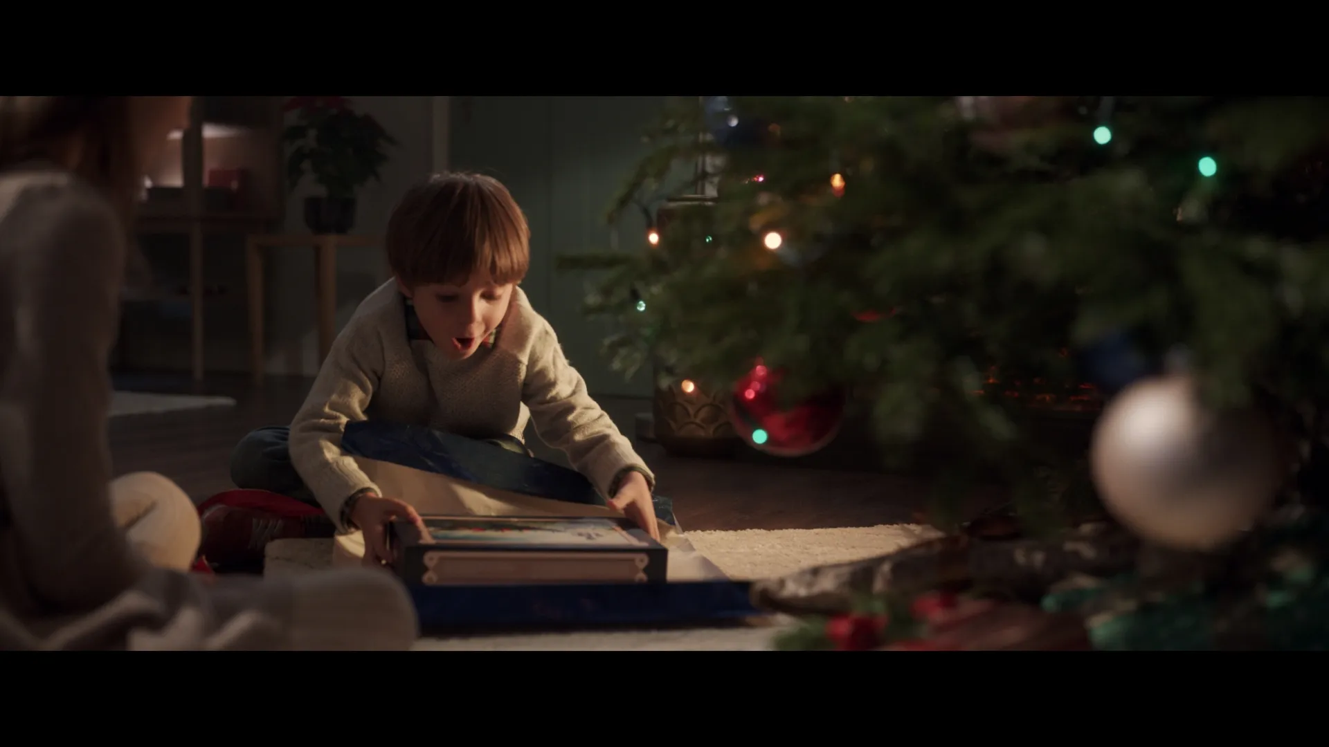 A young boy opens a Christmas present on the floor, his face lighting up with excitement beside the glowing tree.