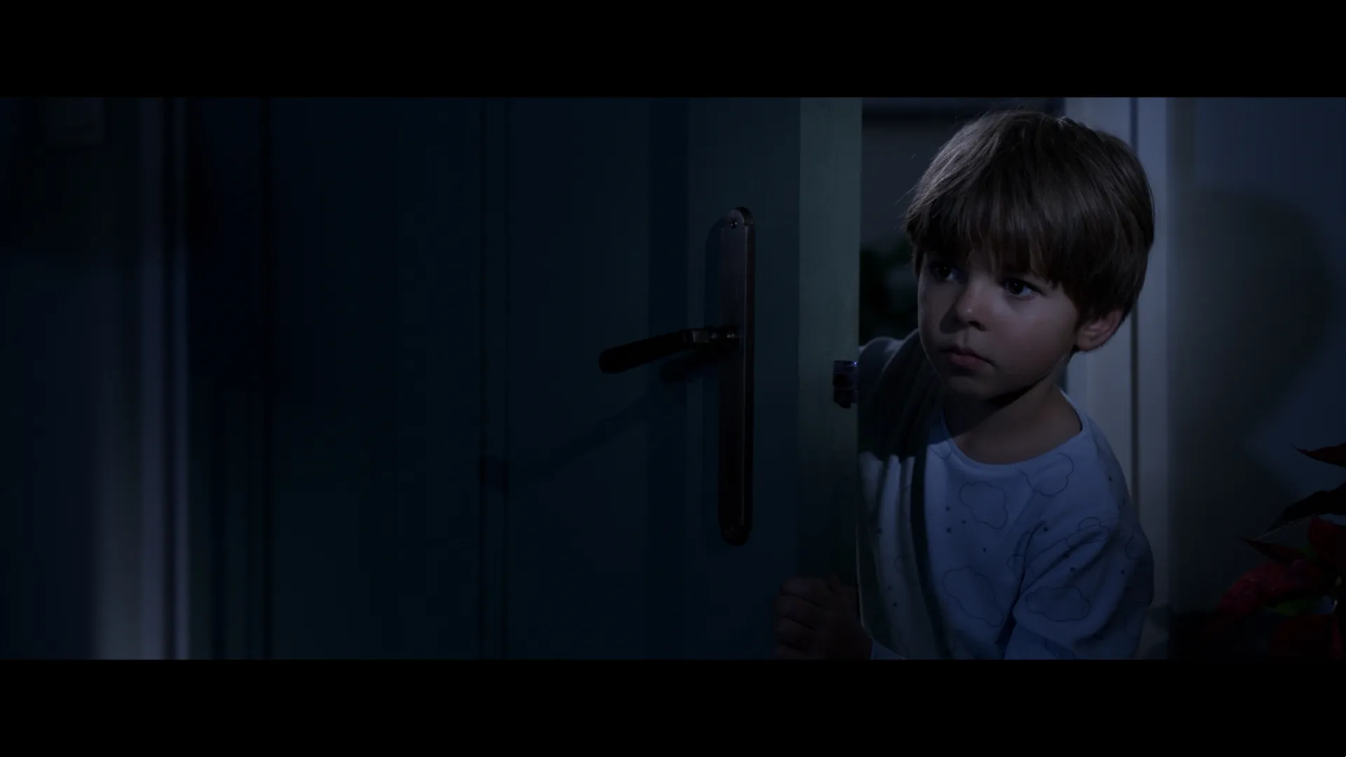 A young boy in pajamas peeks through a cracked door at night, lit softly by moonlight.