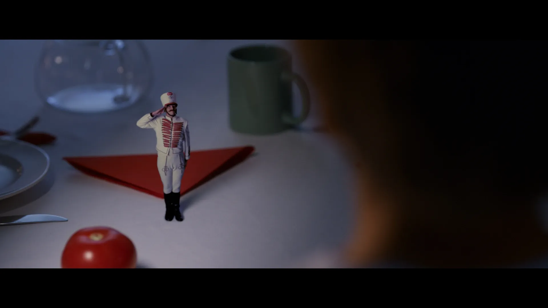 A tiny toy soldier stands saluting on a breakfast table, surrounded by dishes and a folded red napkin.