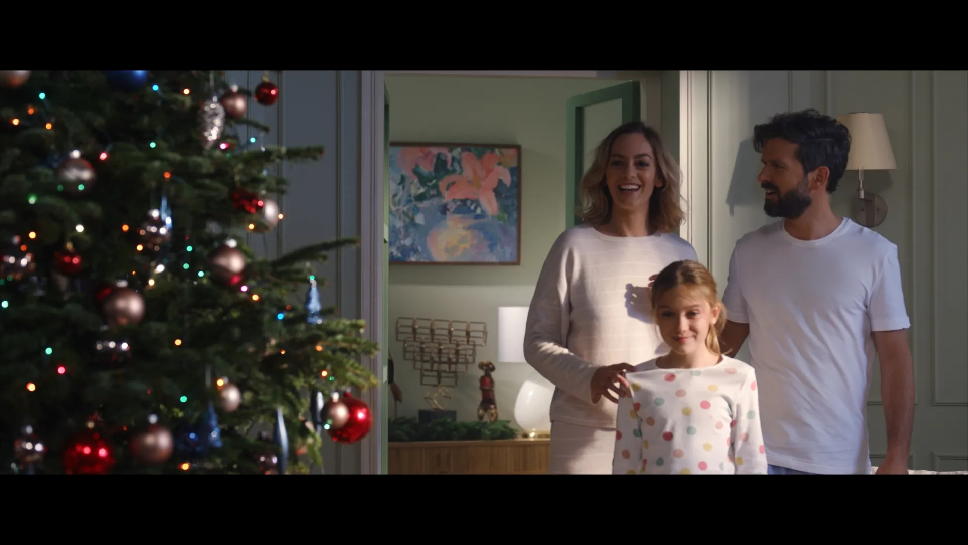 A smiling family steps into the living room on Christmas morning, admiring the lit and decorated tree.