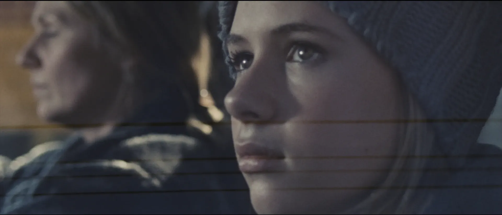 Close-up of a young girl in a winter hat gazing into the distance while an adult sits blurred in the background.