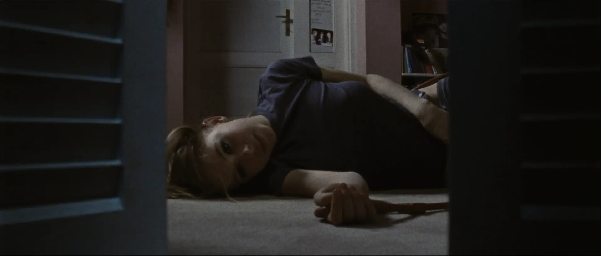 A girl lies on the floor of a dimly lit room, viewed through a narrow gap as she stares ahead with a faint, uneasy expression.