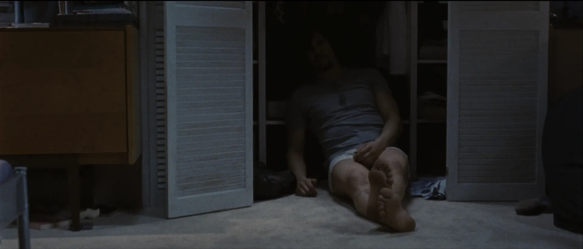 A man sits slumped on the floor inside an open closet, his legs stretched out into the dimly lit room.