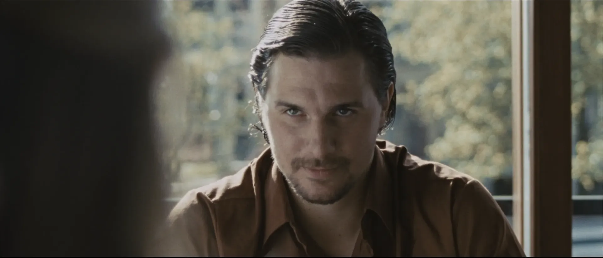 A man with slicked-back hair leans forward, giving an intense, unsettling stare in a softly lit room.