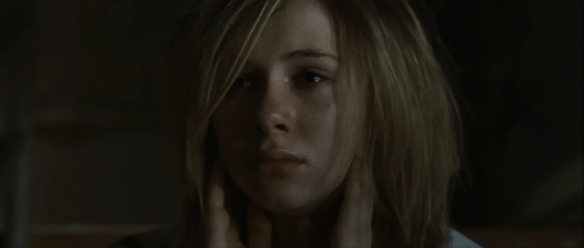 A young girl sits in dim light with her hands at her neck, her expression tense and weary.