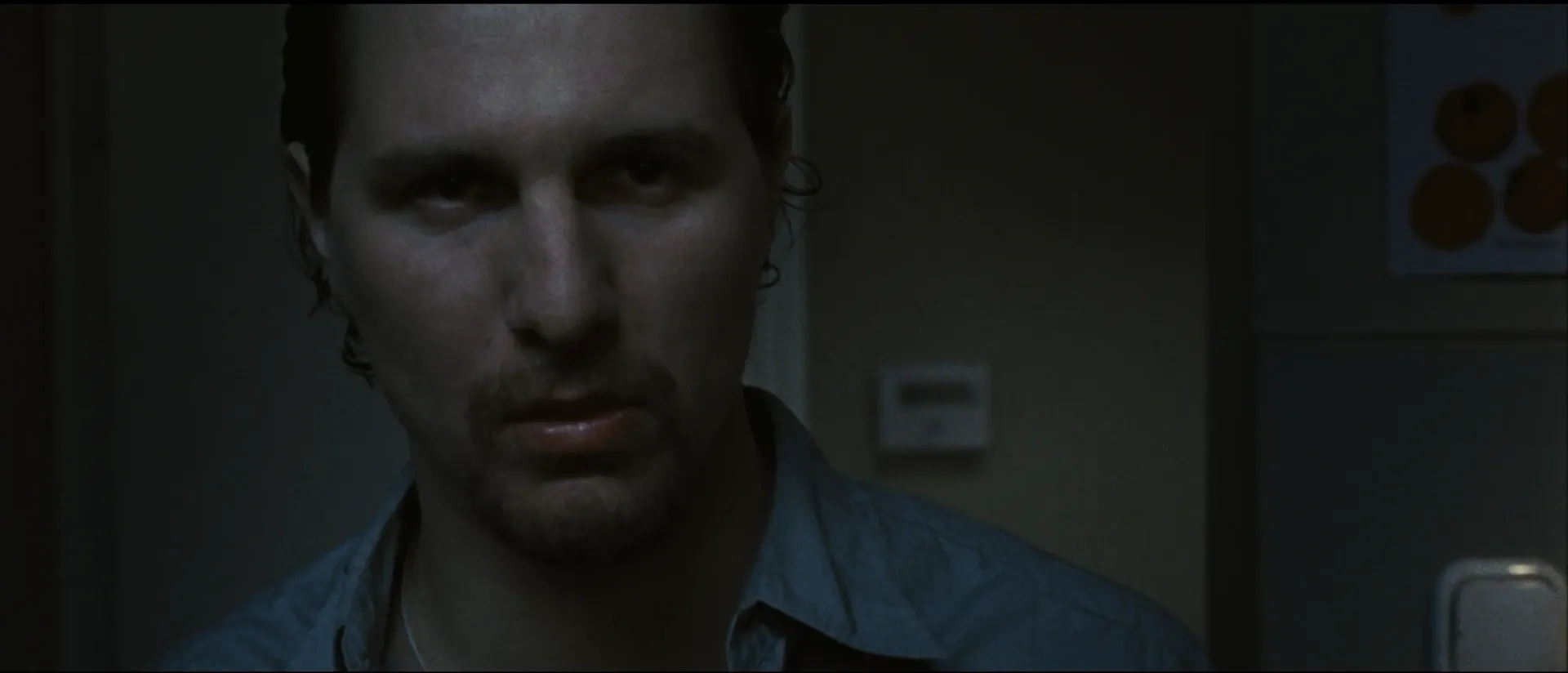 A man stands in dim blue light with a hard, menacing stare, his face partially shadowed.