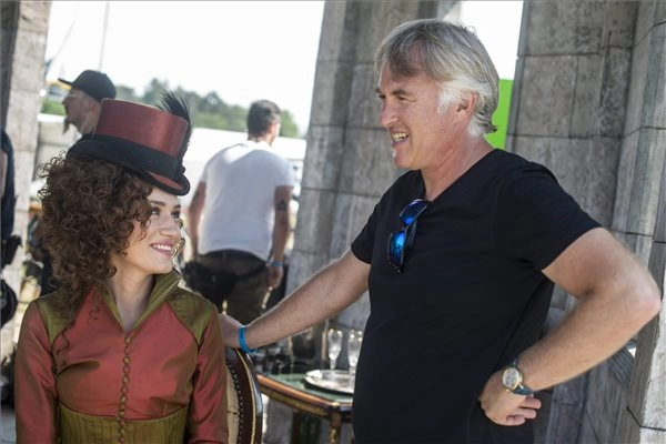 Actress in an ornate red period costume talking with a crew member on set, standing under bright flash lighting.
