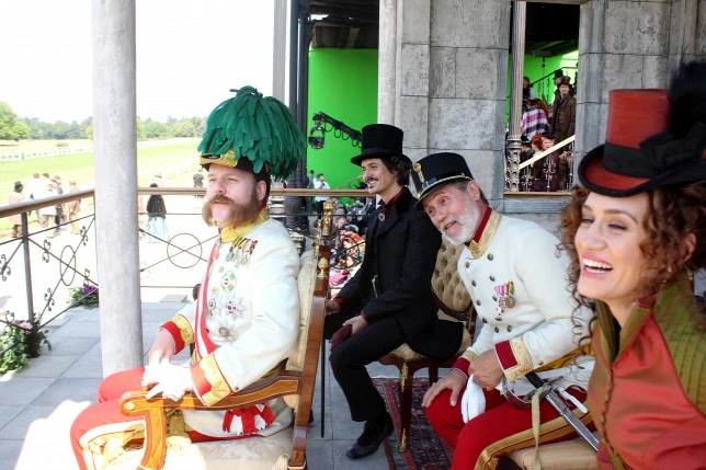 Actors in ornate period costumes sitting in chairs on an outdoor set, smiling and preparing for a scene, photographed with bright flash lighting.