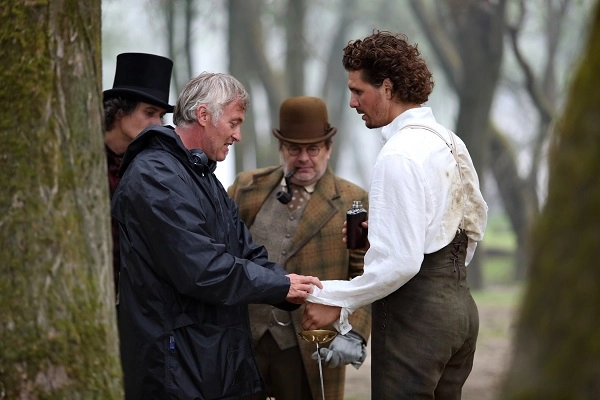 Group of men in period clothing gathered in a forest setting discussing a scene, with flash lighting highlighting their costumes and expressions.