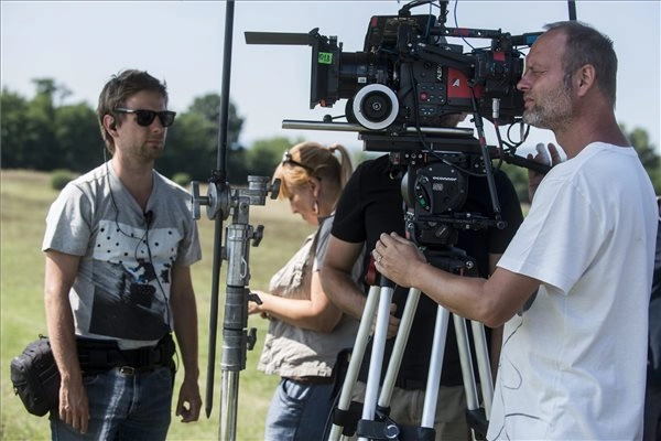 Film crew outdoors operating a professional camera on a tripod, adjusting equipment in bright sunlight enhanced by flash lighting.