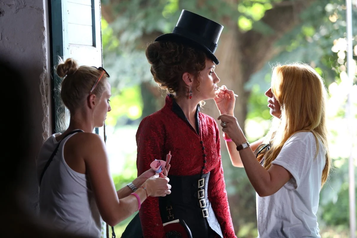 Actress in a red period costume and tall hat being prepared by two makeup artists near a doorway, photographed with bright flash lighting highlighting the details.