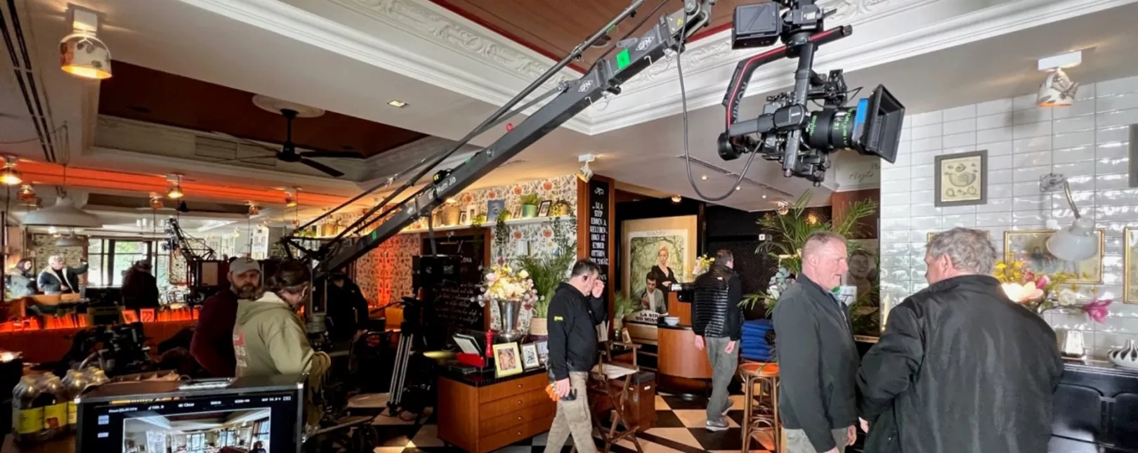 Film crew working inside a decorated café with large camera cranes, lighting equipment, and monitors set up around the space, photographed with flash lighting.
