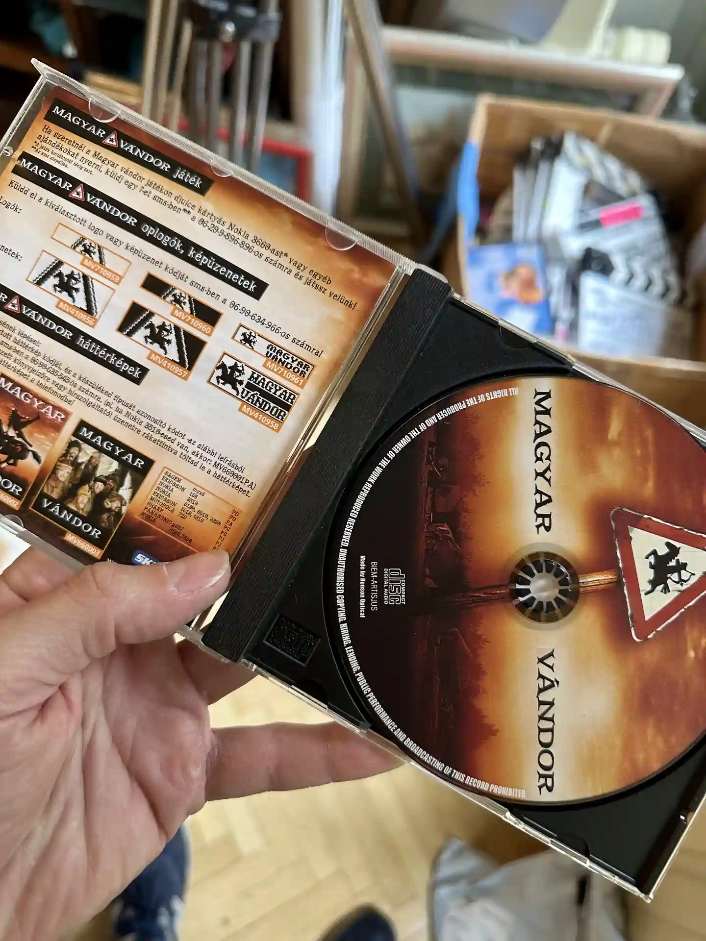 A hand holds an open DVD case for the film Magyar Vándor, showing the disc on the right and printed booklet artwork on the left, with shelves and boxes visible in the background.