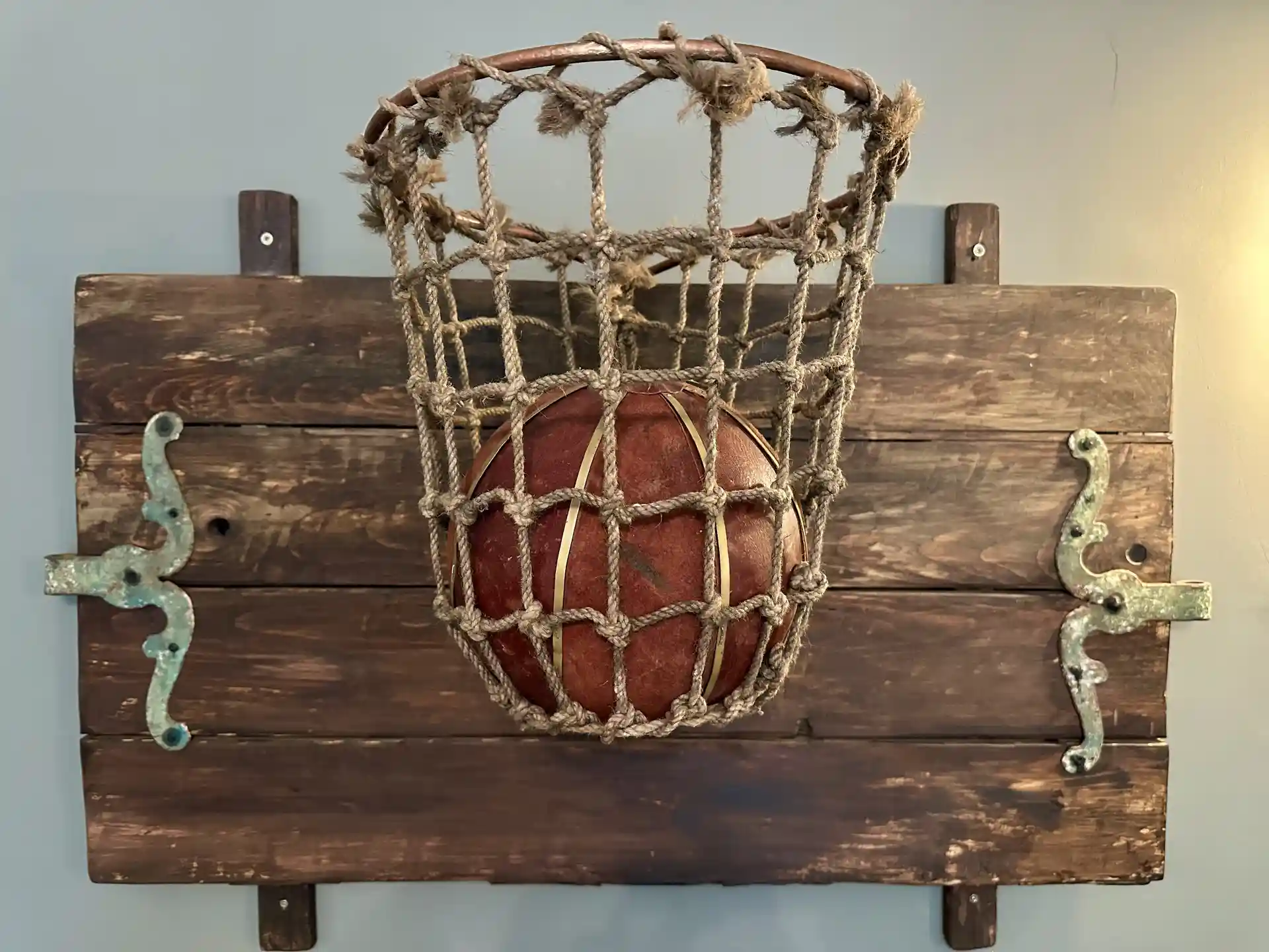 A rustic wooden backboard with a rope basketball net holding an old-fashioned leather basketball, decorated with two small metal horse-and-rider figures on each side.
