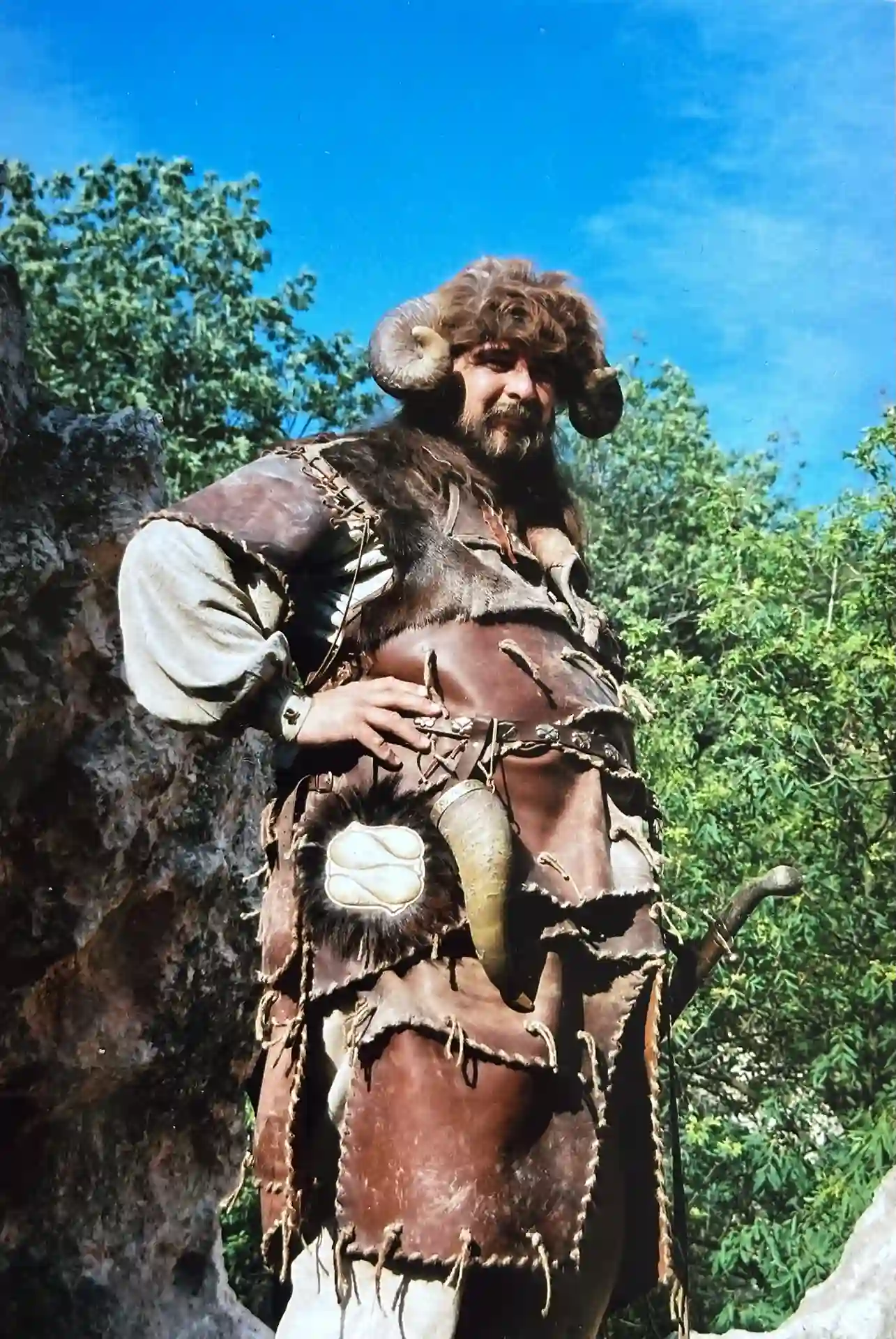 A man in a fur-trimmed, horned warrior costume stands outdoors against a bright blue sky and green foliage, hands on his hips in a proud pose.