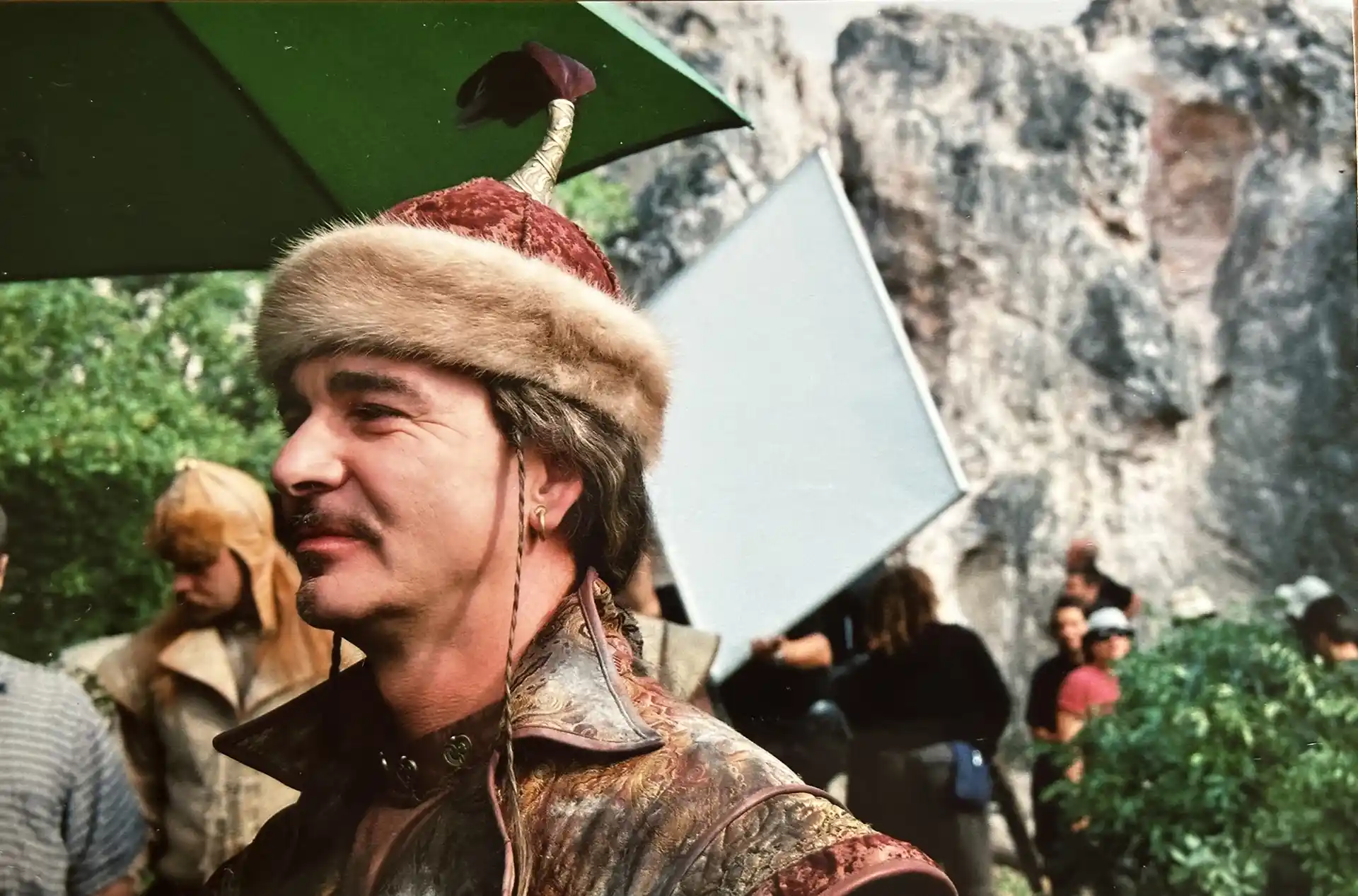 A man in a fur-trimmed hat and medieval-style costume stands outdoors on a film set, surrounded by crew members, greenery, and large reflector panels.