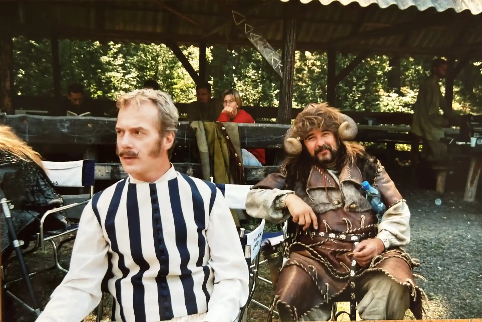 Two men sitting outdoors behind the scenes; one wears a modern striped shirt, while the other is dressed in a fur-trimmed, leather warrior costume with a horned hat.