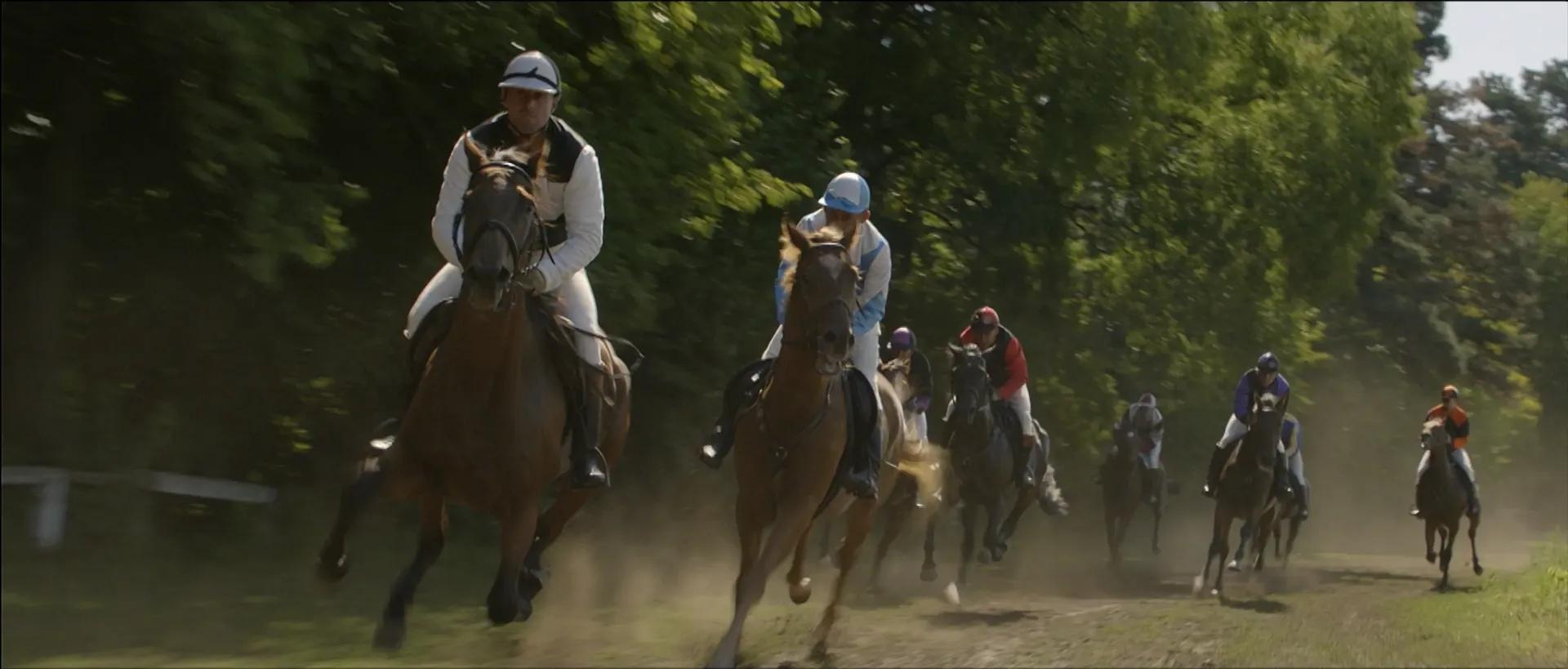 Jokeys gallopping next to one another on a grassy lane next to trees.