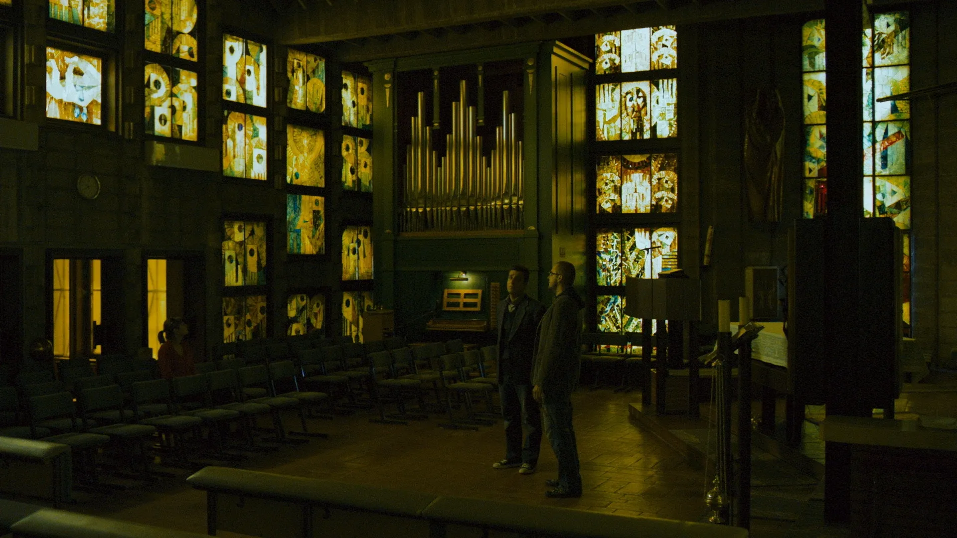 Two people standing in a dimly lit hall with glowing stained-glass windows.