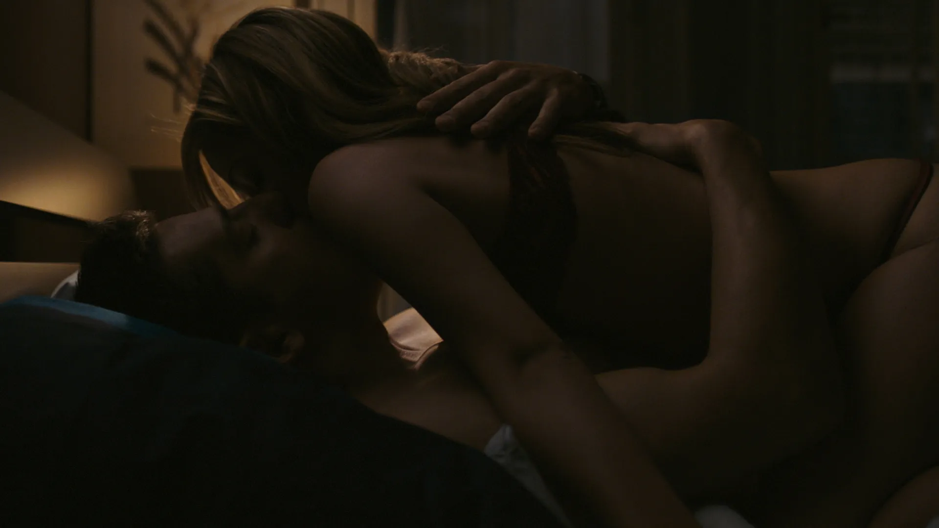 Two actors kissing on a bed in a softly lit bedroom scene from a romantic film.