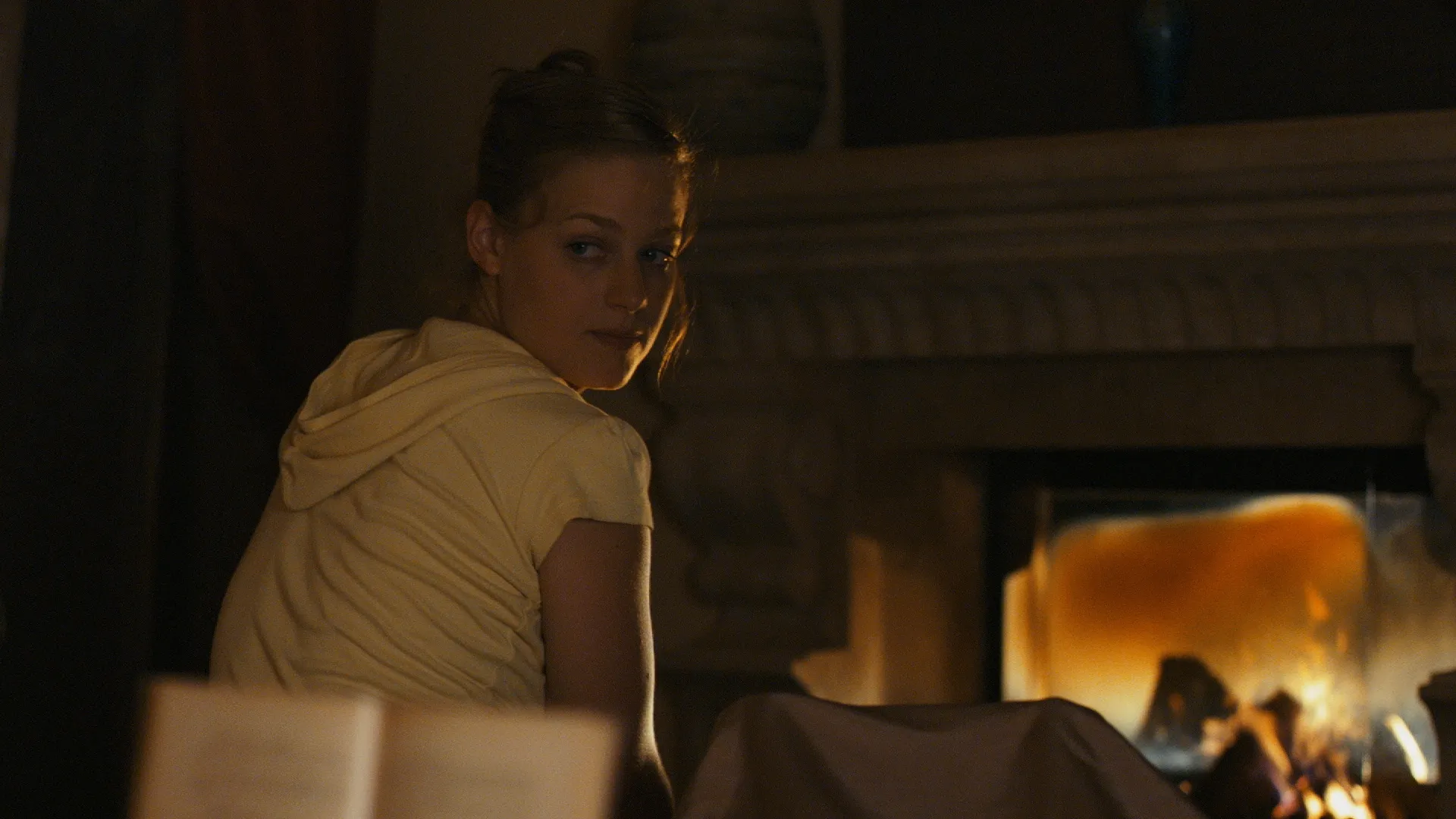 Woman in warm firelight turning back toward the camera with a concerned expression.