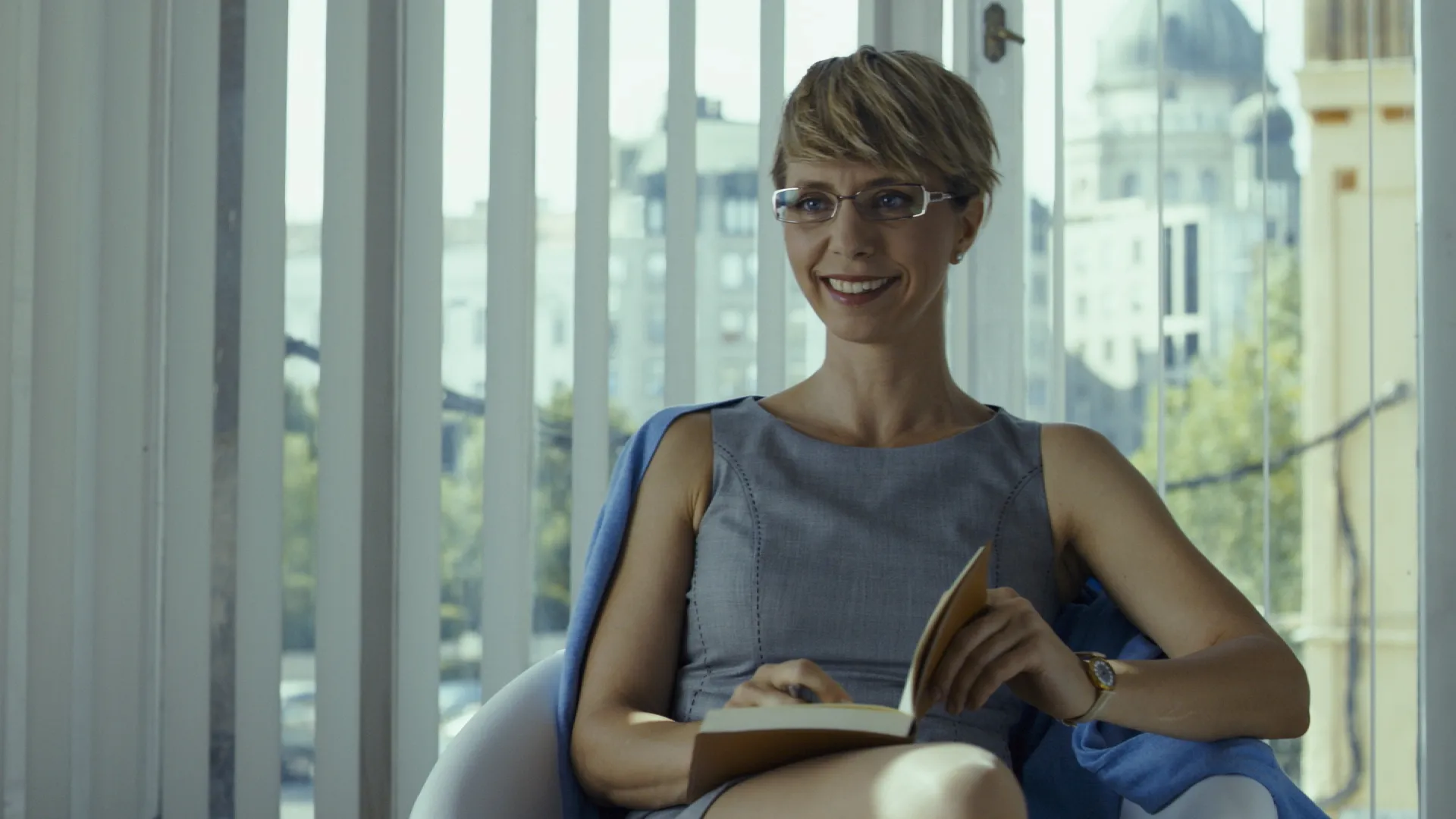 Smiling woman sitting in a bright room, holding an open book on her lap.