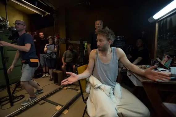 A behind-the-scenes moment on a film set, showing a casually dressed actor gesturing playfully while crew members and cameras prepare around him.