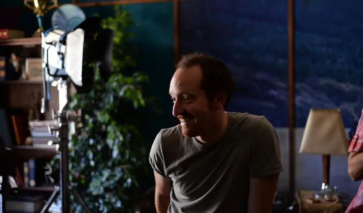 A candid behind-the-scenes moment of a man laughing on set, surrounded by props and warm stage lighting.