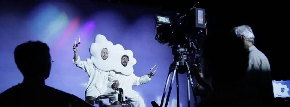 Two performers in oversized white costumes dancing on a brightly lit stage while a camera operator films them in front of colorful lights.