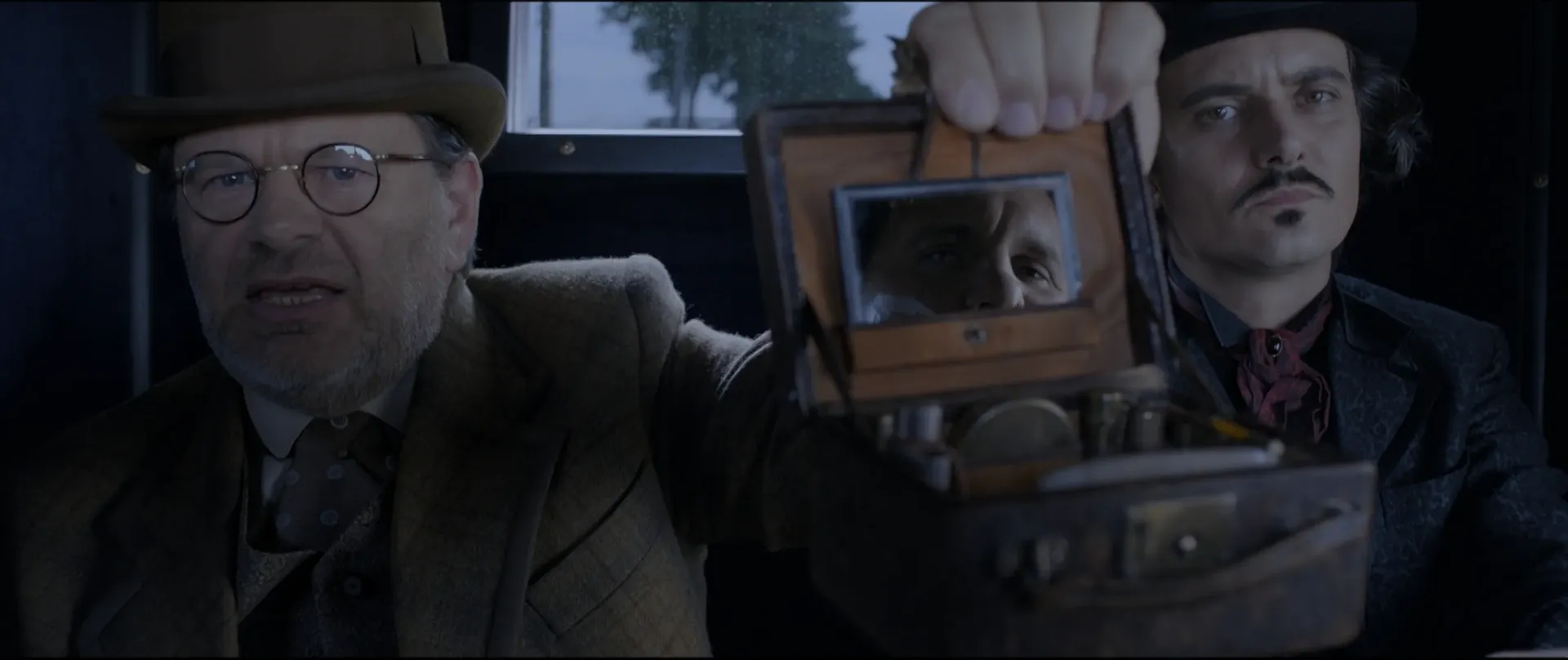 Two men staring intently into the camera one of them is holding up a pocket mirror in which the eyes of third person are shown.