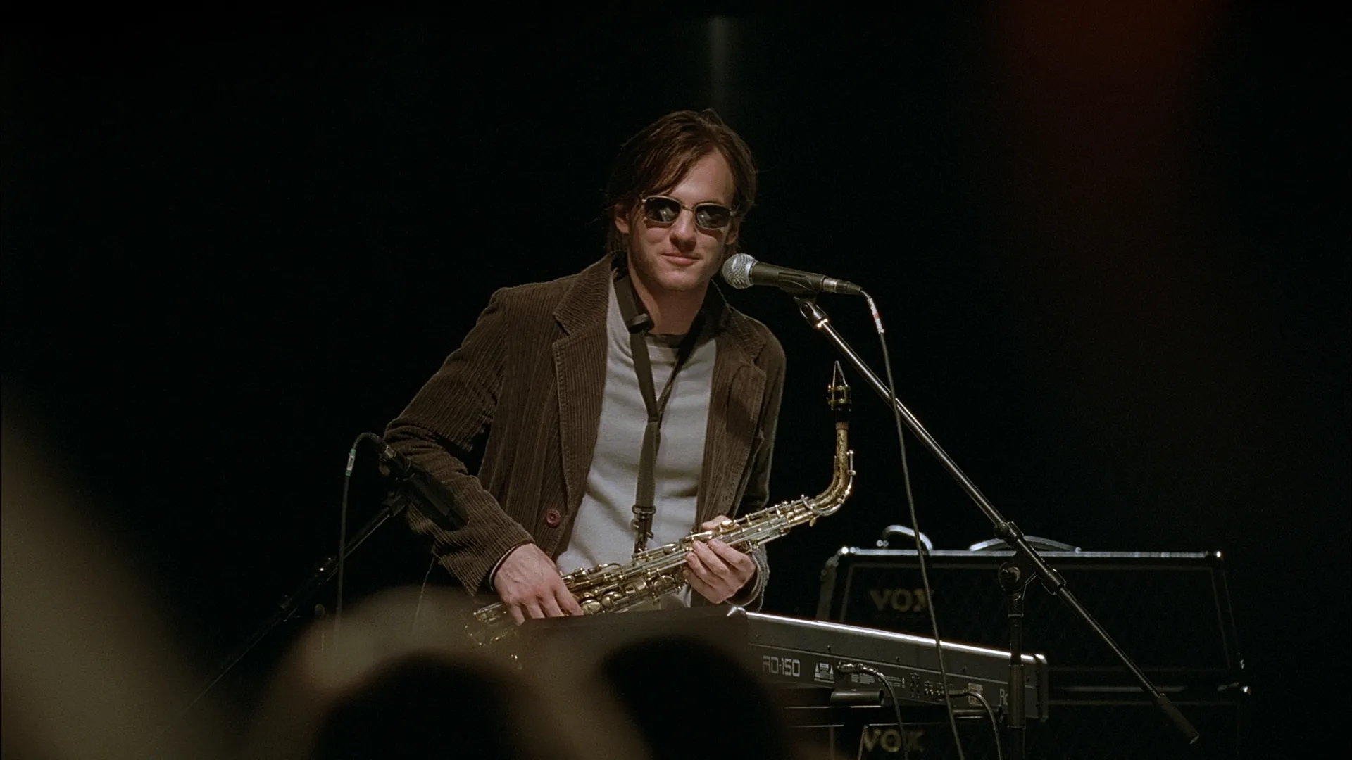A musician wearing sunglasses performs on stage, playing a saxophone and standing behind a keyboard, with the dimly lit background highlighting the live concert atmosphere.
