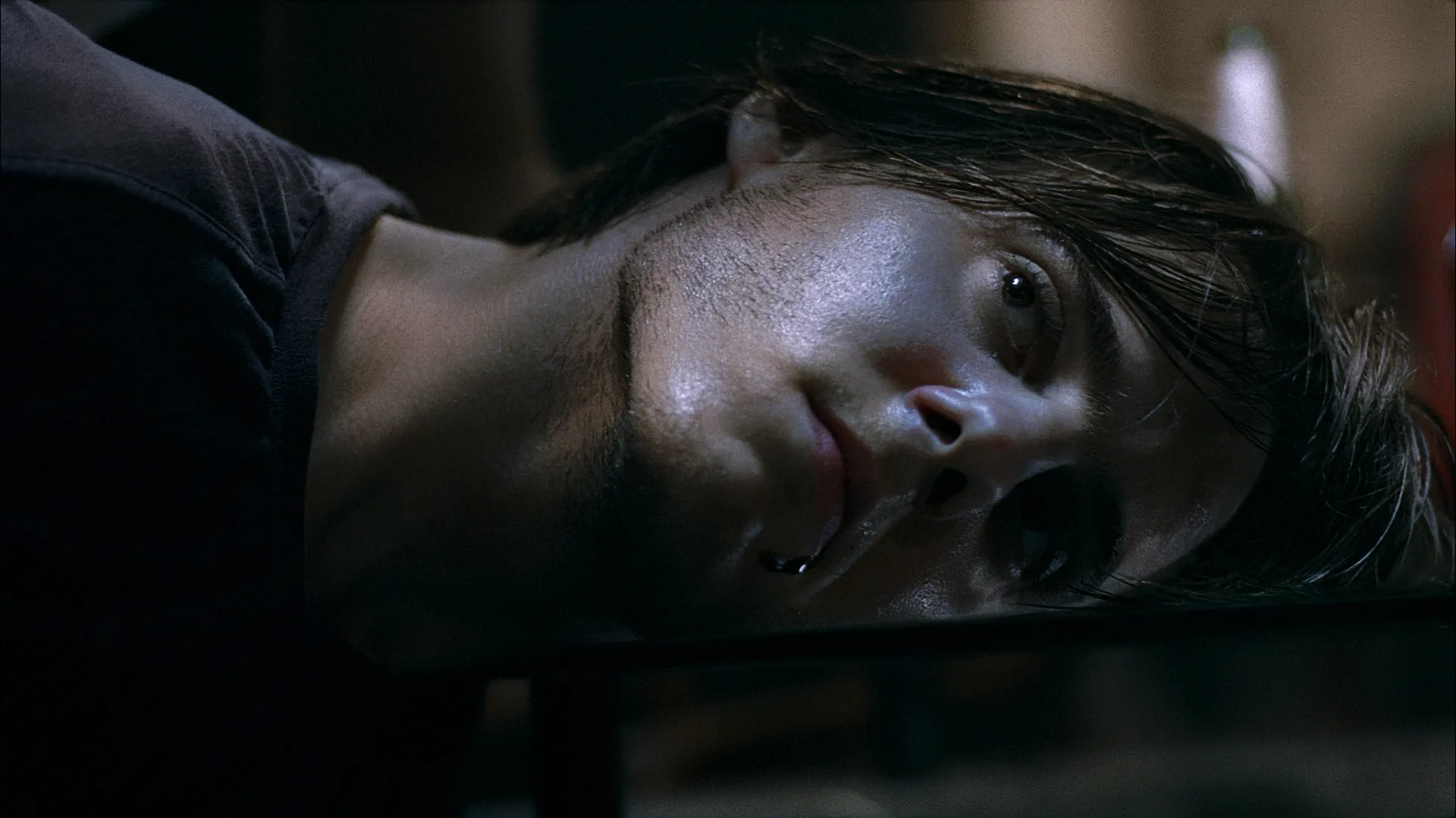 A woman lies on her side in dim, cold lighting, her face pale and expression vacant as she stares ahead.