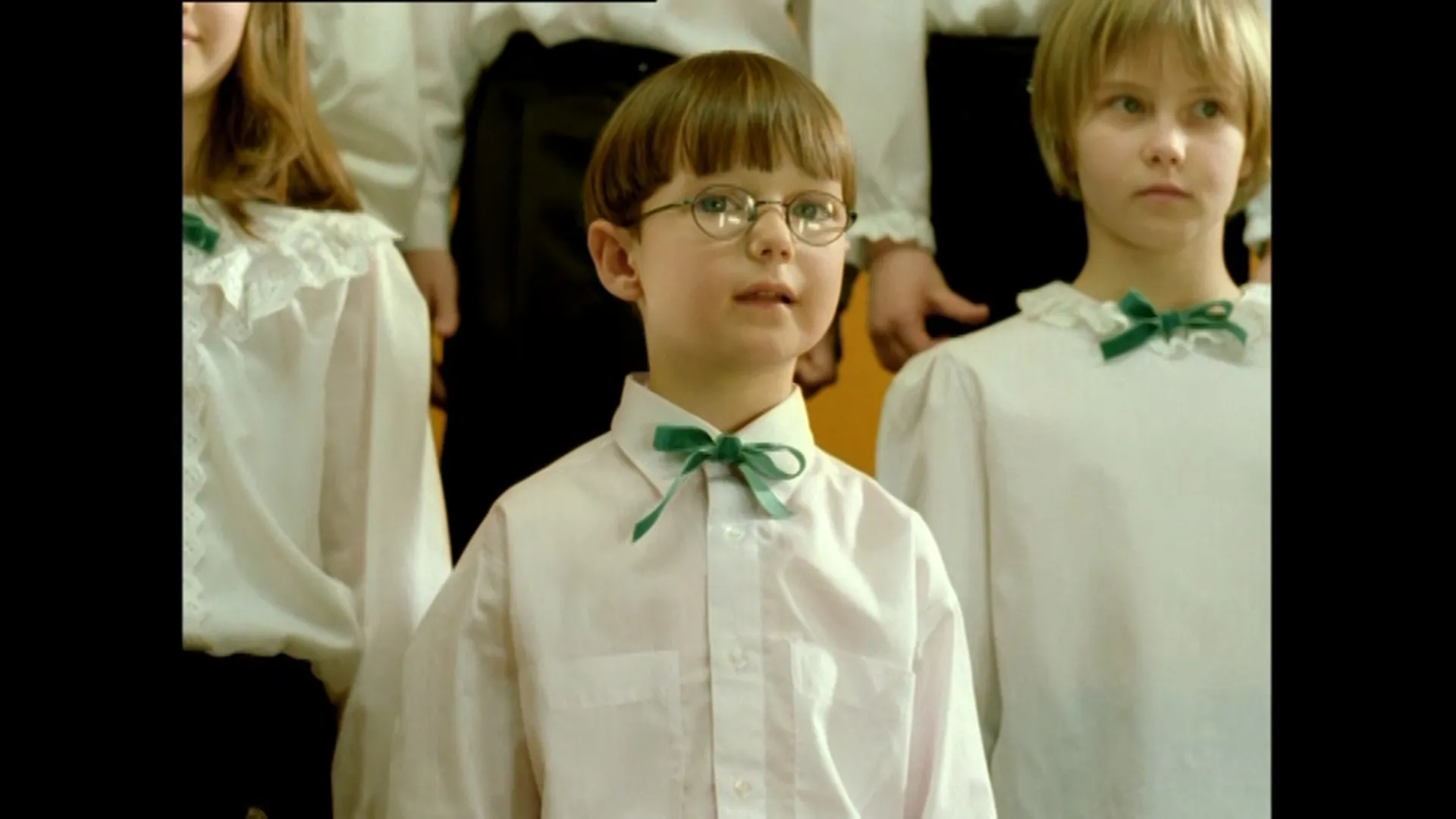 A young boy in glasses stands among children in matching white outfits with green bows, looking slightly upward during a choir-style scene.