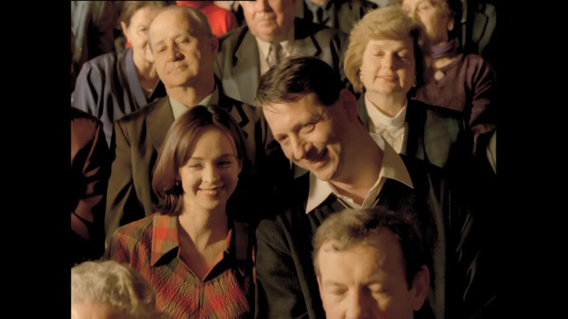 A warmly lit audience sits closely together, with a smiling couple in the center surrounded by people watching attentively.