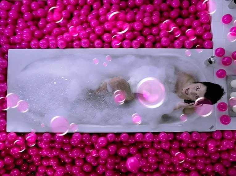 Woman relaxing in a bubble bath, surrounded by bright pink plastic balls while talking on the phone.