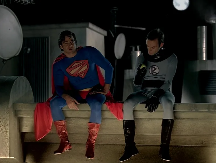 Two men in superhero costumes sit on a rooftop ledge at night, talking under the moonlight.
