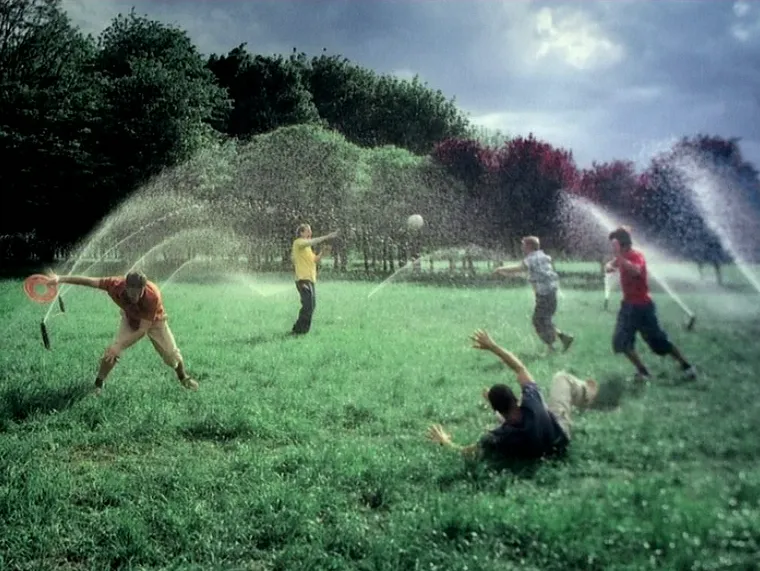 People playing ball on a grassy field while arcing sprinkler streams spray across the scene.