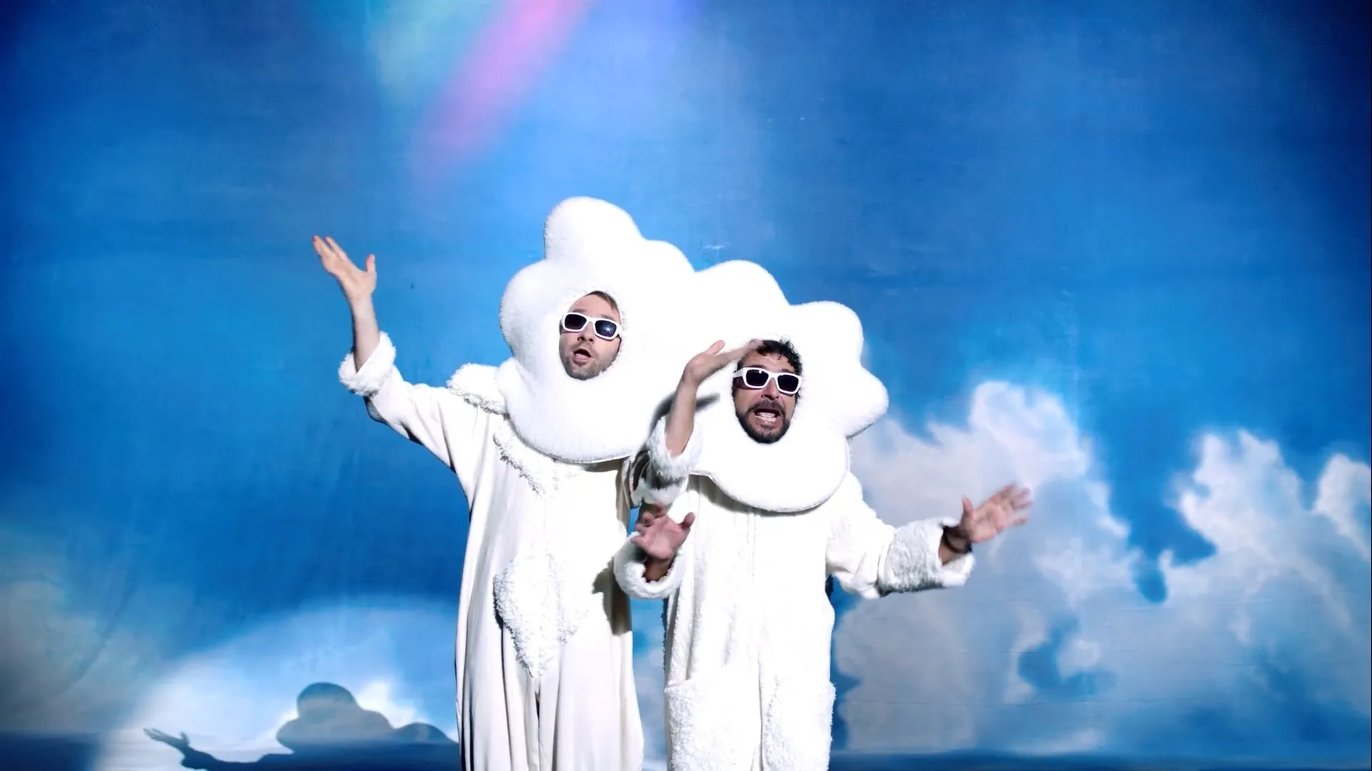 Two performers dressed as fluffy white clouds, wearing matching sunglasses, enthusiastically gesture against a bright sky-themed backdrop.