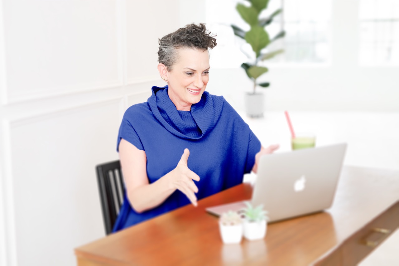 Color photo of Tessa smiling and talking to laptop while sitting in a chair at a table