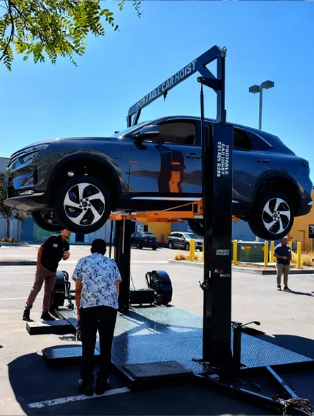 A blue SUV is elevated on a portable car hoist outdoors with three people nearby inspecting it.