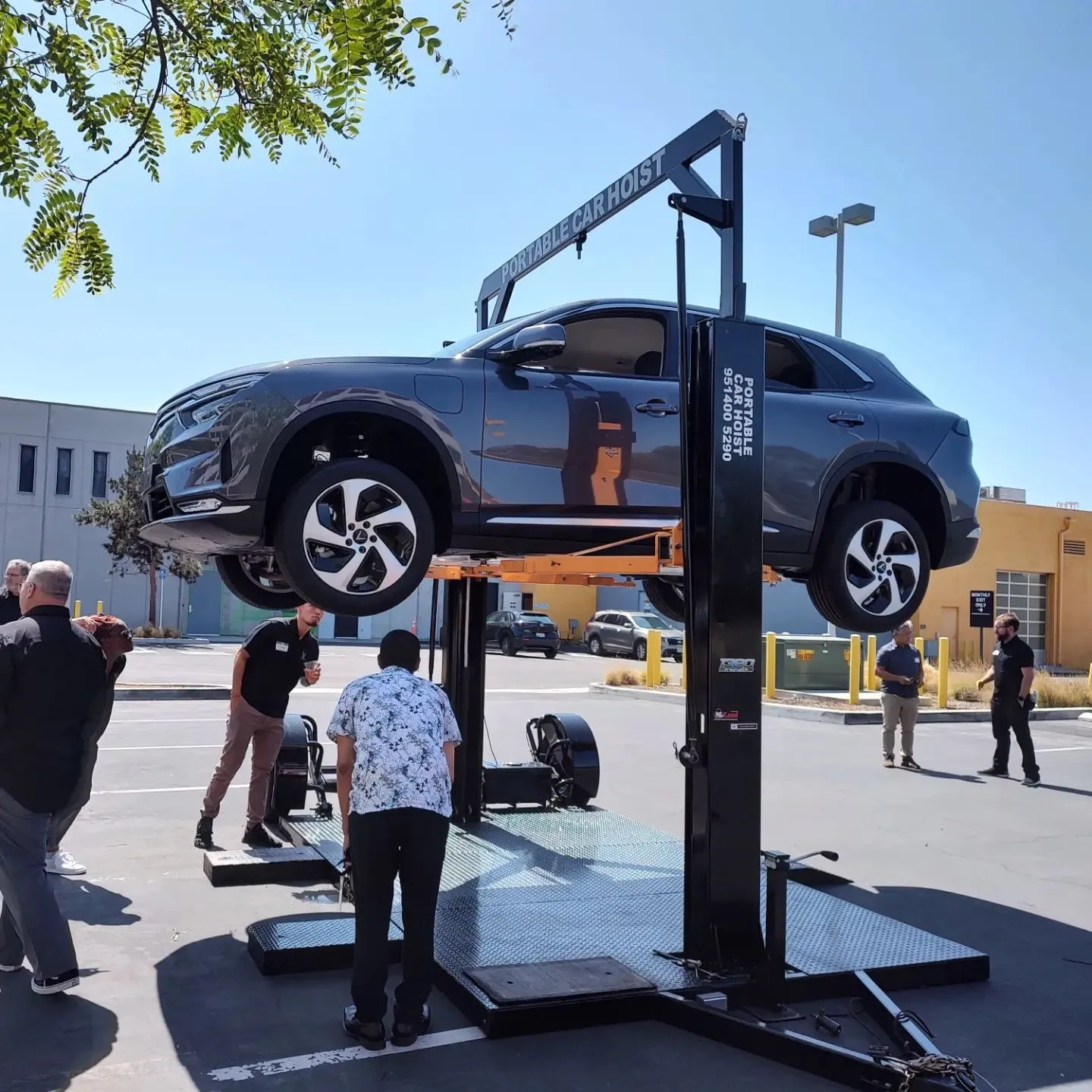 Gray SUV lifted on a portable car hoist with people observing and discussing outdoors.