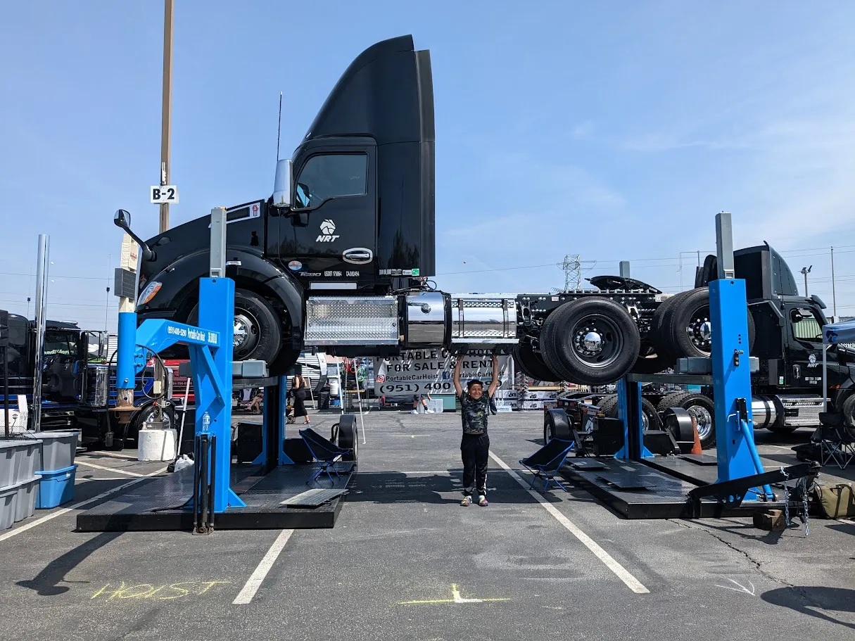 Black semi-truck raised on blue hydraulic lifts with a child standing underneath holding it up.