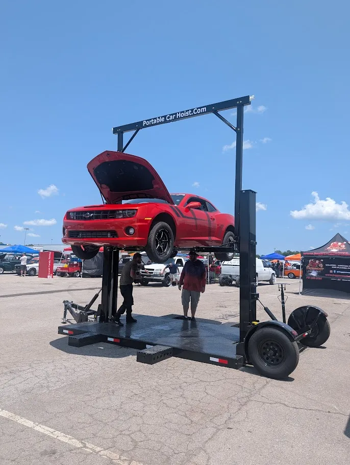 Red car lifted by a portable car hoist at an outdoor event with two people nearby.