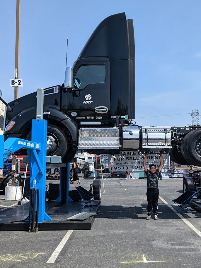 Person standing beneath a lifted black semi-truck on a blue car hoist in a parking lot.