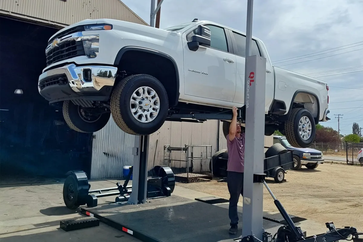 Man working underneath a white Chevy pickup truck lifted on a portable car hoist outside a garage.