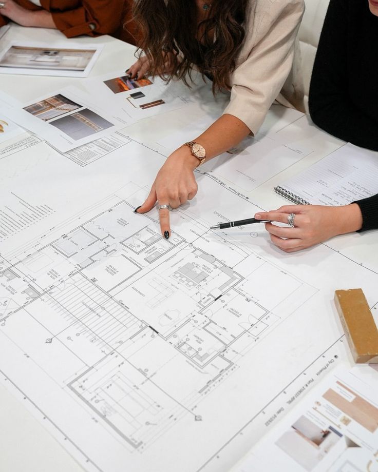 Two people reviewing architectural floor plans spread on a table, one pointing with a finger and the other holding a pen above the drawings.